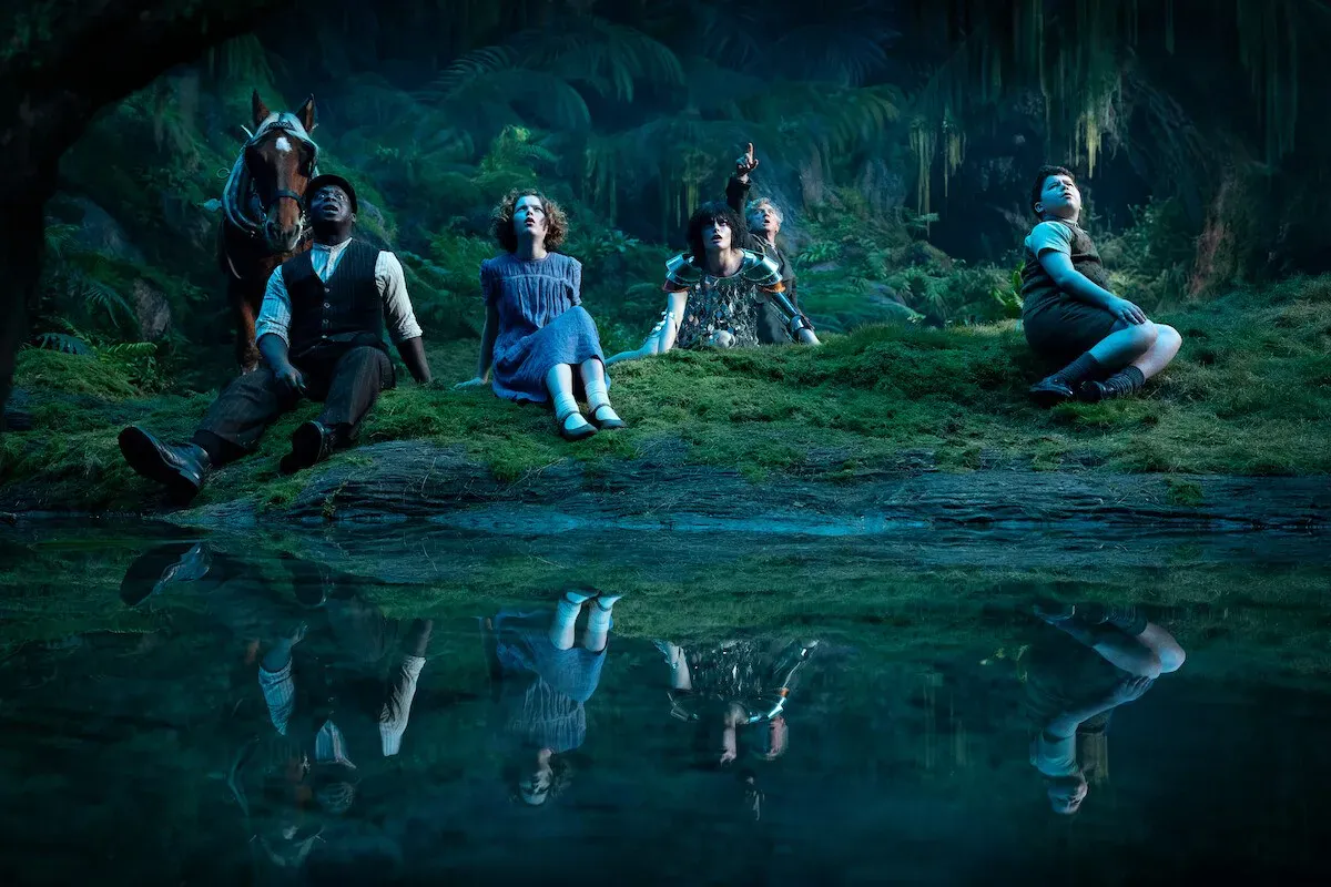 Grupo de personagens sentados na grama à beira de um lago noturno em cena de Nárnia: O Sobrinho do Mago