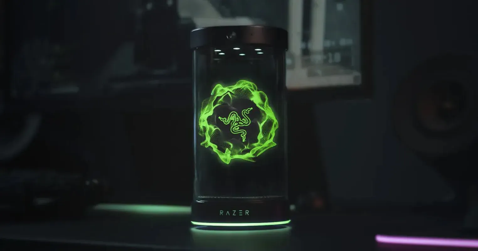 Dispositivo cilíndrico Razer AVA com o logotipo verde da Razer em holograma, sobre uma mesa escura