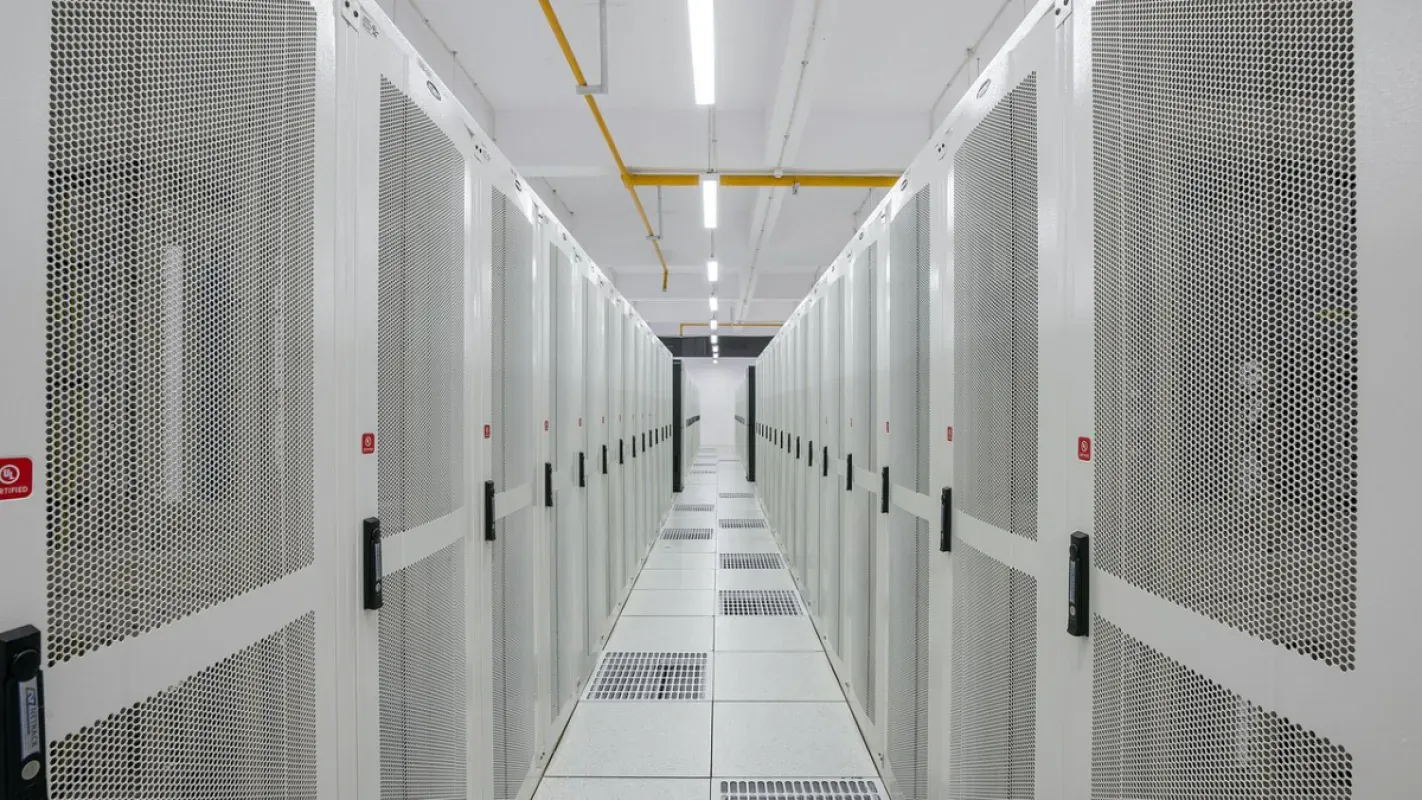 Corredor entre duas fileiras de racks brancos fechados em um data center, com piso elevado e luzes no teto